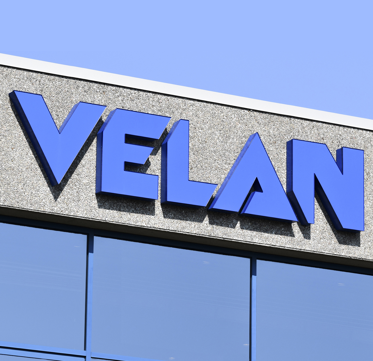 The image shows a building with a large blue sign displaying the word "VELAN" against a textured gray surface. Below the sign, there are windows reflecting a clear blue sky.