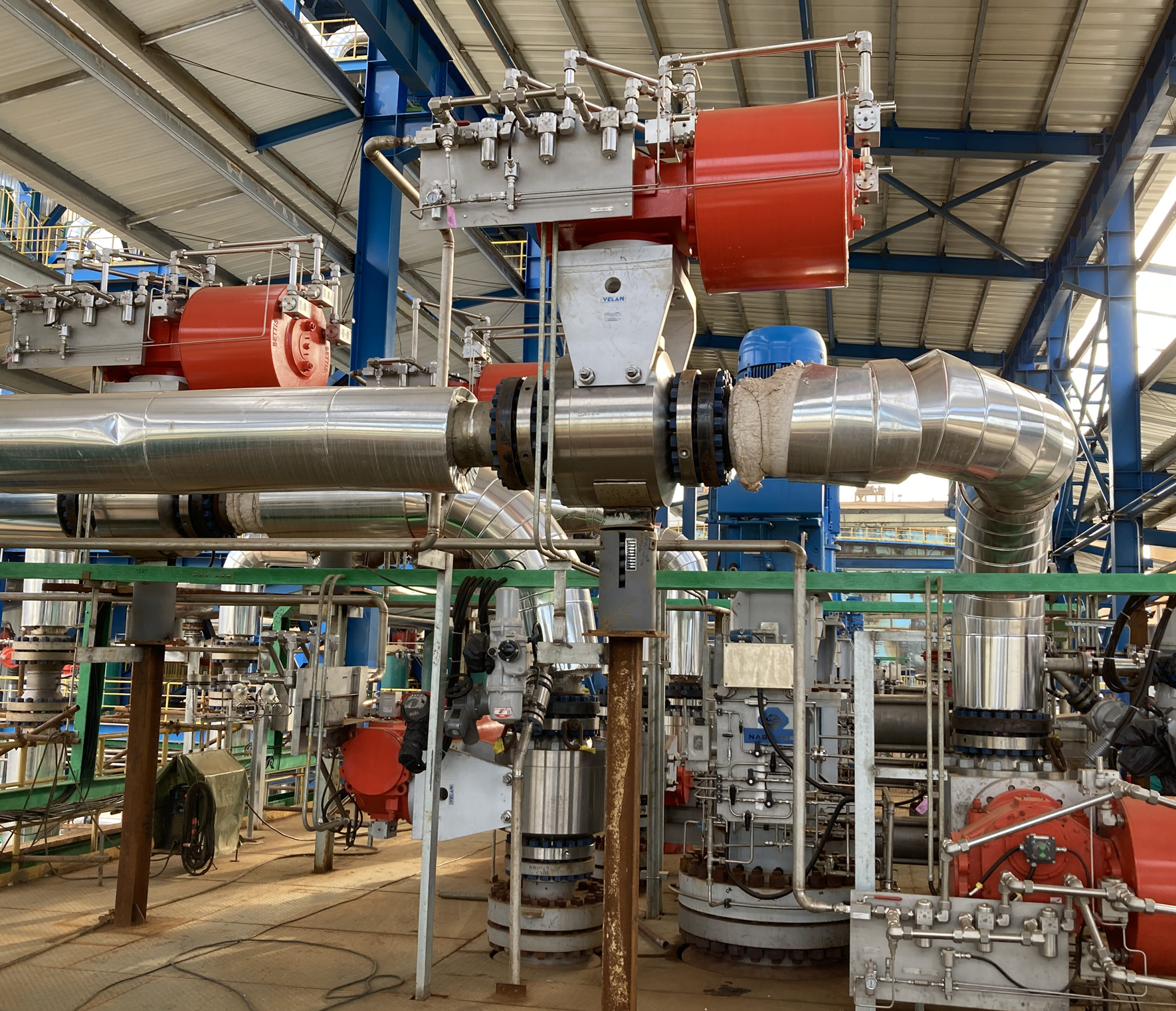 A large industrial facility with interconnected metallic pipes and machinery. The structure is supported by blue steel beams, and red mechanical components are visible among the pipes. The floor appears unfinished with some equipment placed around.